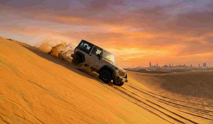 A 4x4 vehicle is depicted driving up a steep sand dune at sunset, kicking up sand, with a city skyline in the distance, illustrating an off-road adventure where following Dune bashing safety tips Saudi Arabia is crucial.