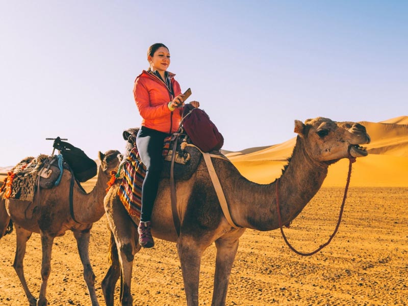 A guide in traditional white thobe and red-and-white ghutra leading two camels through the Jeddah Desert, with a woman in a beige top and a man in a green shirt riding the camels against a backdrop of golden sand dunes and rocky mountains.