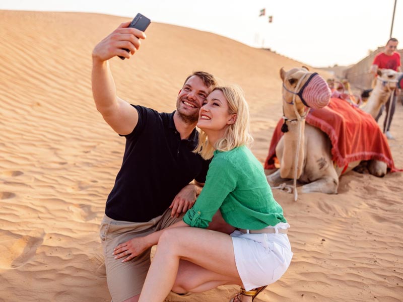 Woman in a red jacket riding a camel while holding a phone, with another camel beside her in the golden sands of the Jeddah Desert, Saudi Arabia.