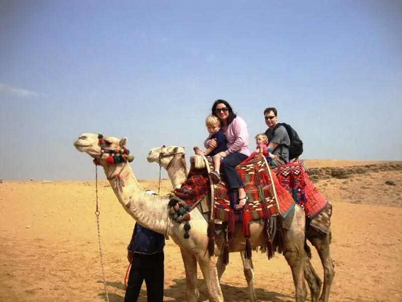 Man in traditional white thobe and red-and-white ghutra standing beside a seated camel in front of a large striped Bedouin tent in the Jeddah Desert.