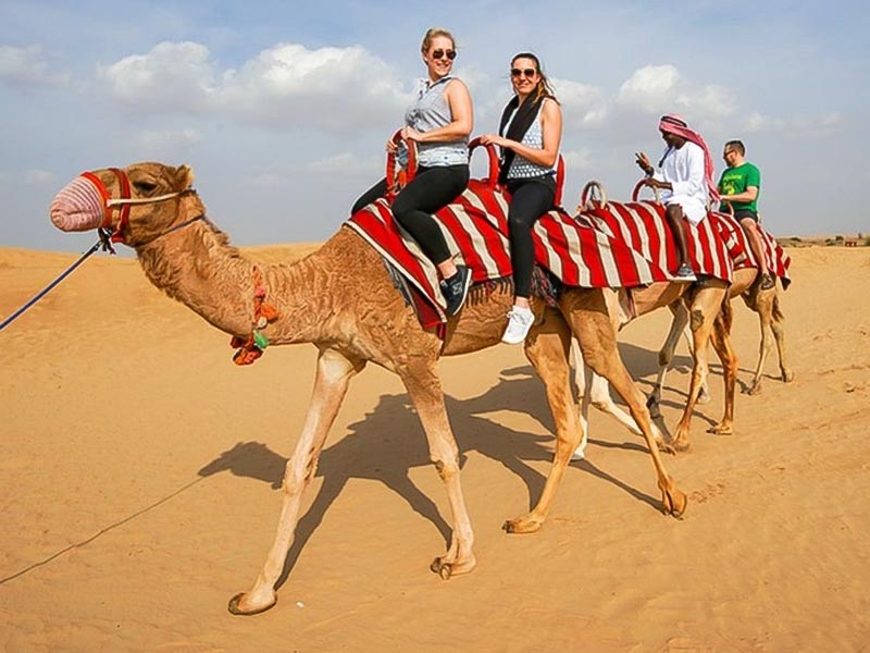Two women smiling while riding camels side-by-side during a guided camel trek in the Jeddah Desert, with their guide visible behind them.