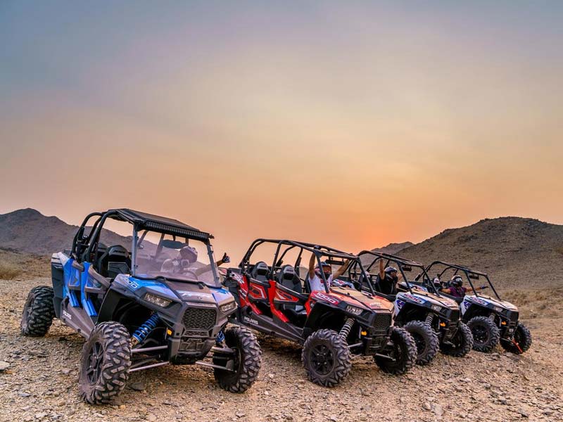 A line of colorful Polaris dune buggies parked in the golden Jeddah desert at sunset, with excited tourists waving from the vehicles during a dune buggy tour.
