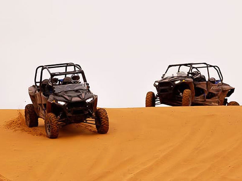 Two black dune buggies speeding over a large sand dune in the Jeddah Desert, kicking up sand as they conquer the terrain on an off-road adventure.