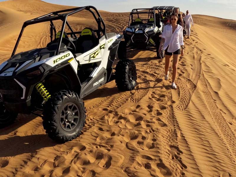 A white and black Polaris RZR dune buggy parked on the sandy tracks of the Jeddah Desert, with a woman in a white shirt walking nearby and more vehicles visible in the background.