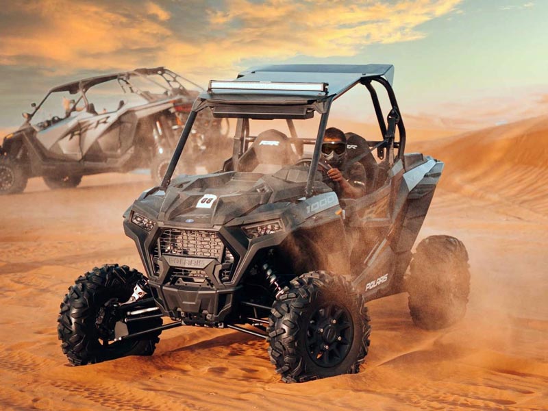 A black Polaris 1000 dune buggy aggressively driving through the Jeddah Desert sand, creating a large dust cloud, with another buggy visible in the background during a thrilling dune tour.