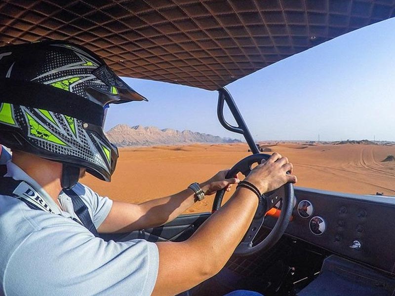 Close-up view from inside a dune buggy in the Jeddah Desert, showing a driver wearing a helmet and gripping the steering wheel while enjoying the vast orange sand dunes and mountain landscape.
