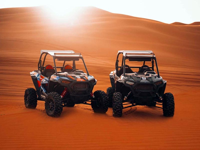 Two Polaris dune buggies parked side by side on the stunning orange sand dunes of the Jeddah Desert, with dramatic sunlight and lens flare creating a beautiful desert backdrop.