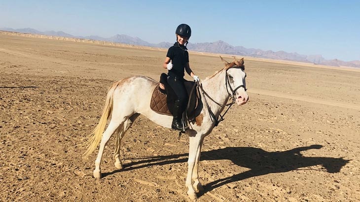 Young rider wearing a helmet giving a thumbs-up while mounted on a light-colored horse in the Jeddah desert.