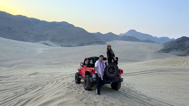A yellow Jeep Wrangler carrying passengers drives through the sandy terrain of the Jeddah desert.