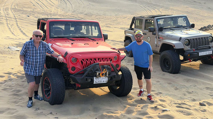 A couple leans against a white SUV at the top of a sand dune, watching the sunset with other safari groups in the background.