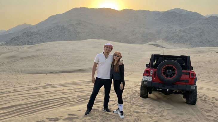 People riding yellow quad bikes over rolling sand dunes during a vibrant orange sunset in the desert.